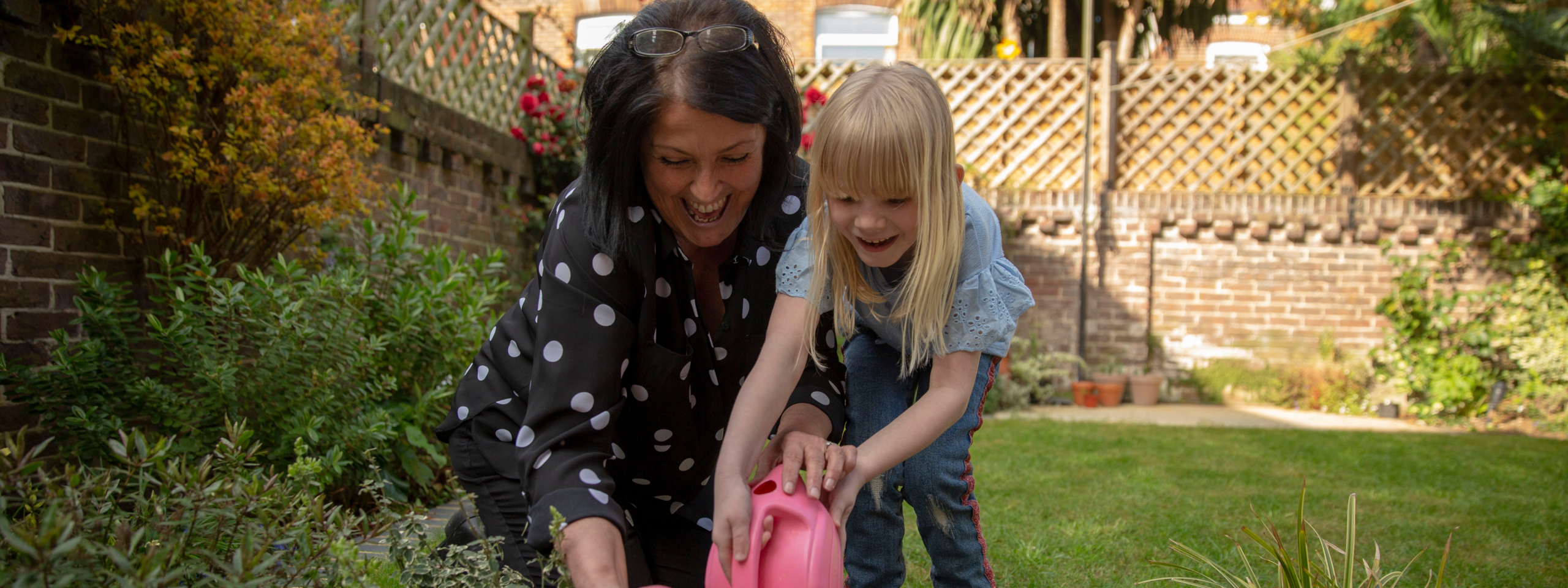 Laughing woman with a young blond girl laughing and watering plants in a garden>